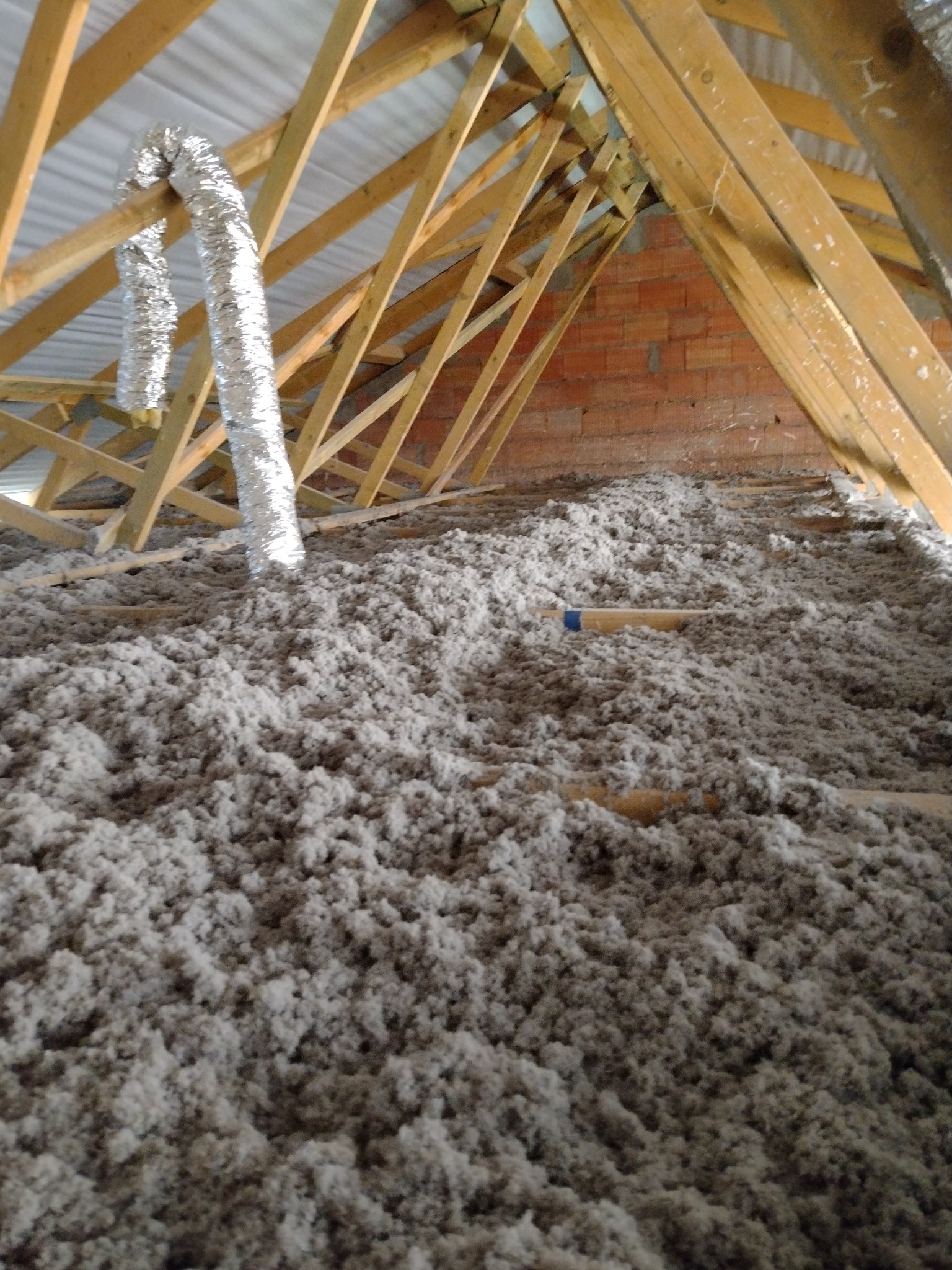 Poddasze z widoczną drewnianą konstrukcją dachu, ocieplone szarą wełną mineralną, z fragmentem aluminiowej rury wentylacyjnej w rogu.