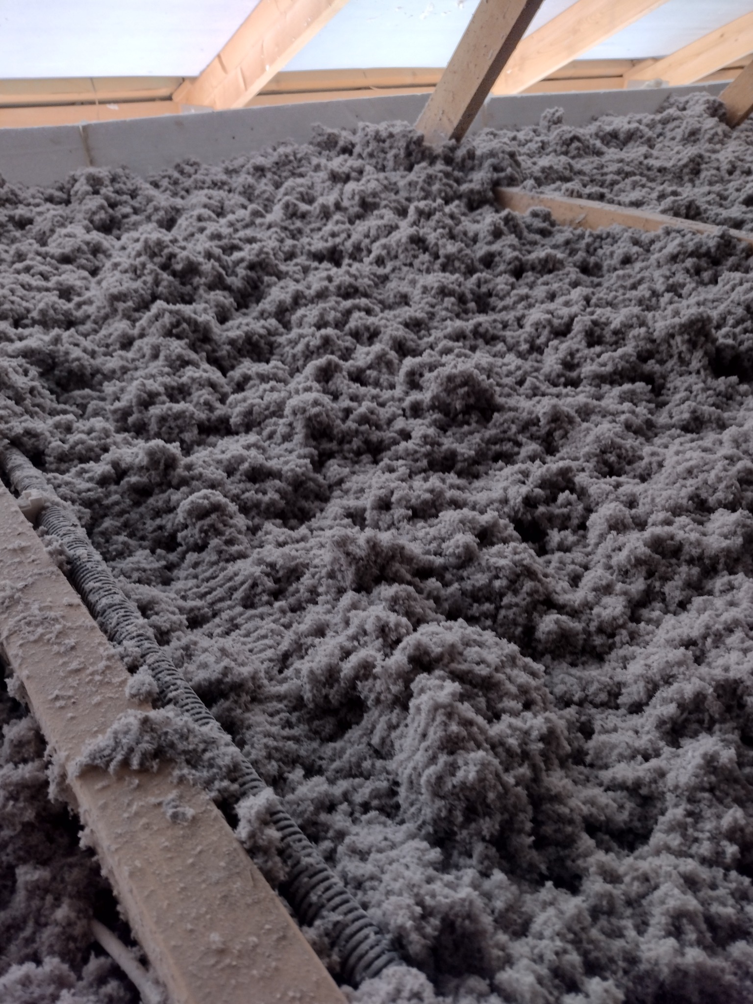 Szara izolacja celulozowa wypełnia przestrzeń poddasza między krokwiami, widoczne przewody instalacyjne i drewniane elementy konstrukcji dachu.