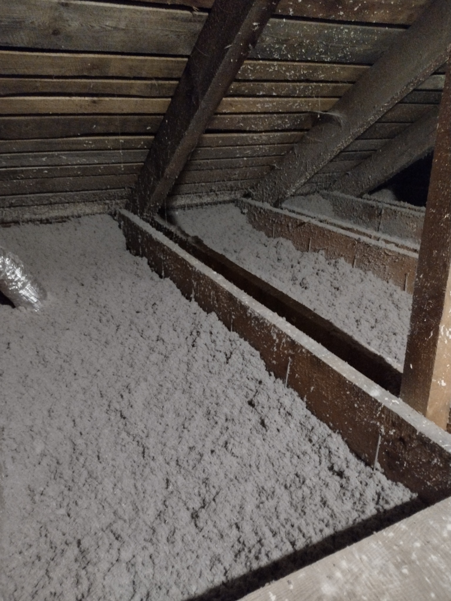 Poddasze z drewnianą konstrukcją wypełnione szarą wełną izolacyjną, widoczny fragment przewodu wentylacyjnego.