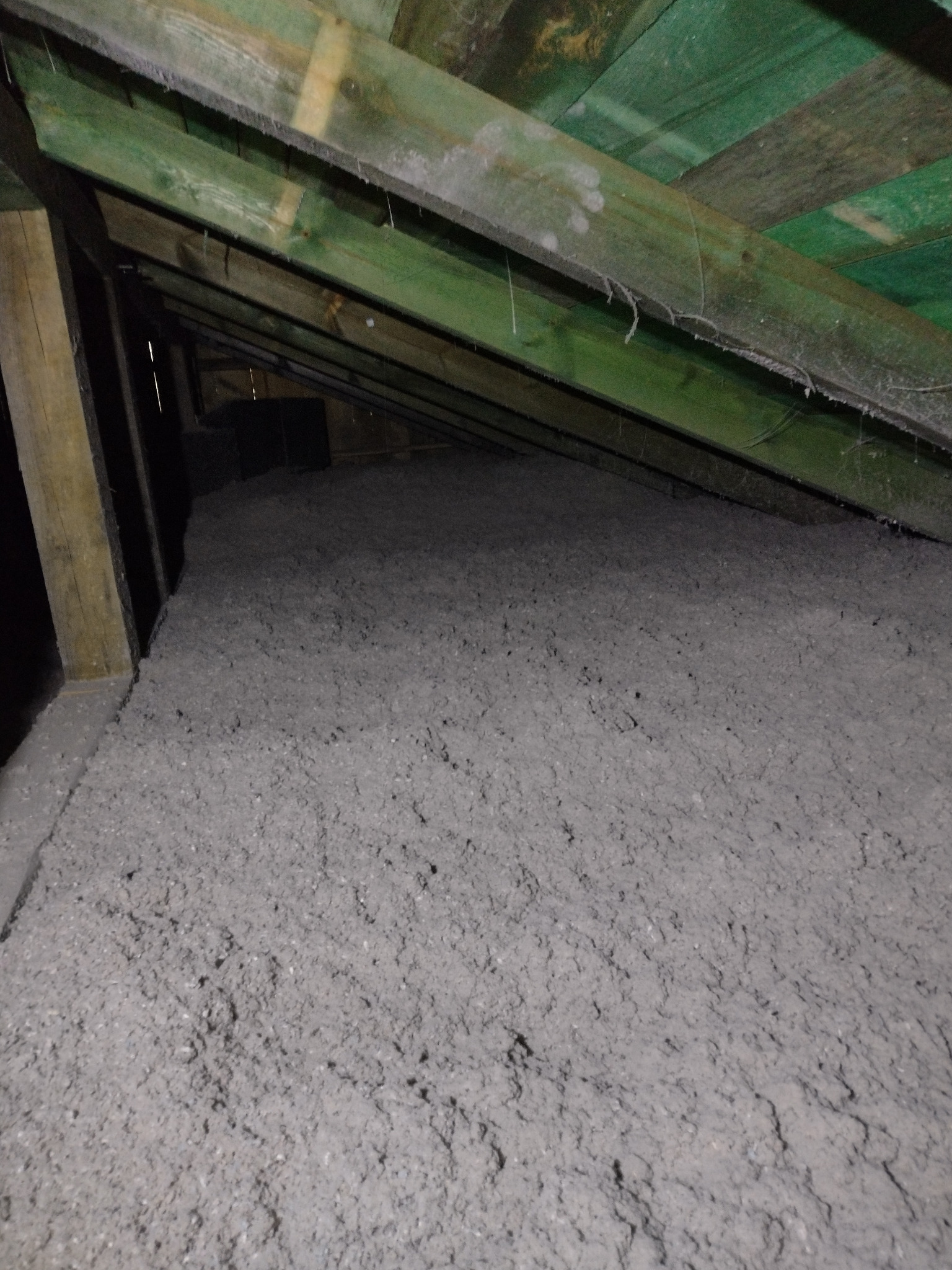 Szara, sypka izolacja termiczna wypełniająca przestrzeń poddasza, widoczne zielone drewniane elementy konstrukcji dachu.