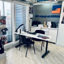 Wnętrze biura firmy MS więcej niż OKNA z ekspozycją okien z żaluzjami poziomymi, biurkiem, krzesłami i materiałami reklamowymi.