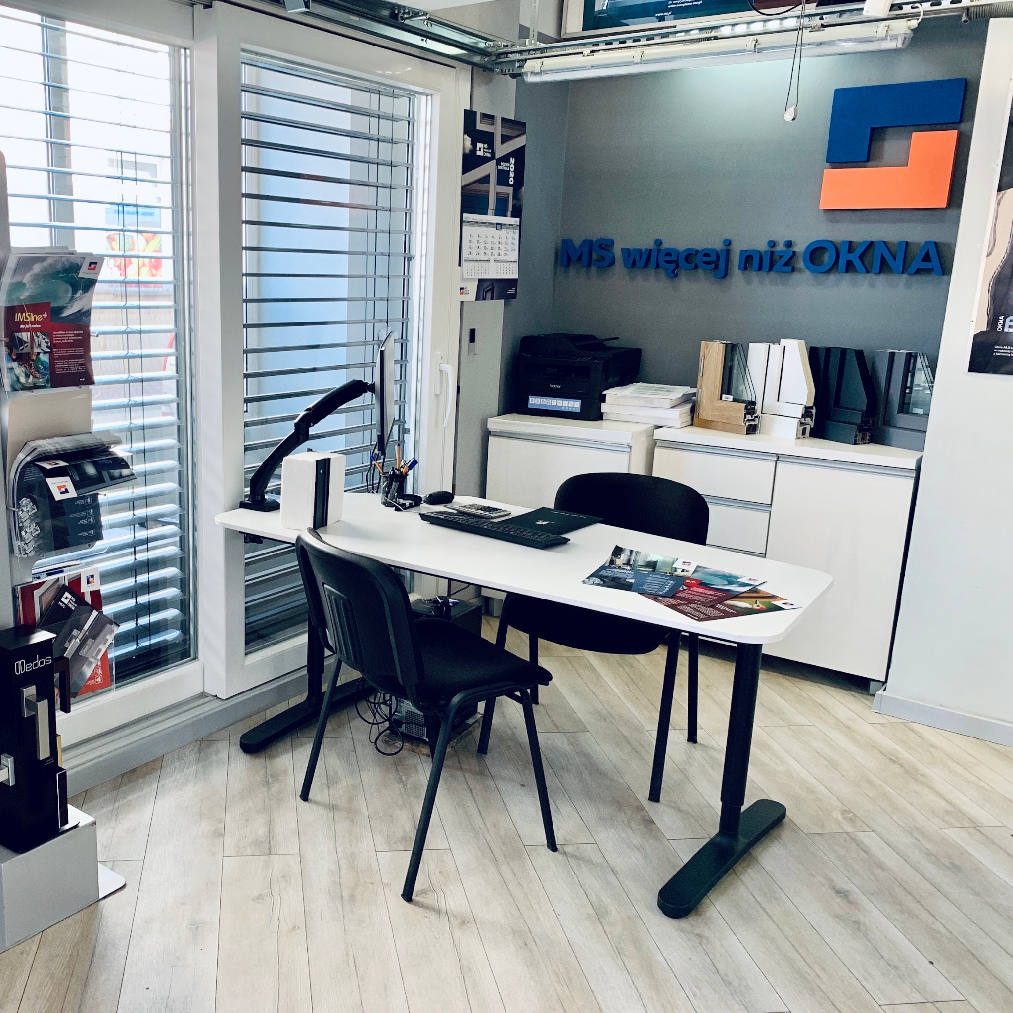 Wnętrze biura firmy MS więcej niż OKNA z ekspozycją okien z żaluzjami poziomymi, biurkiem, krzesłami i materiałami reklamowymi.