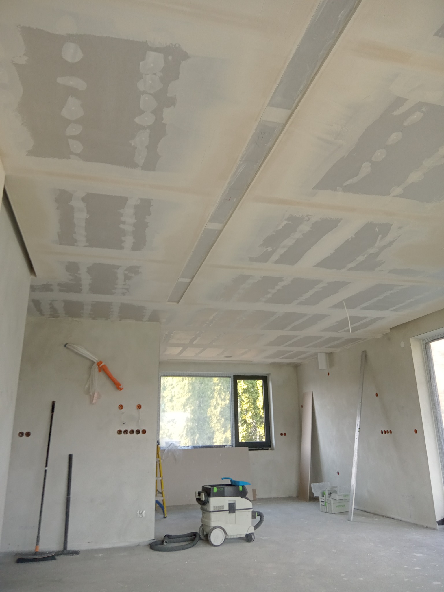 Wnętrze w trakcie remontu, widoczne przygotowane ściany i sufit z widocznymi łączeniami płyt, szpachlowane, ale jeszcze nie pomalowane, na podłodze stoi odkurzacz przemysłowy, a przy ścianie...
