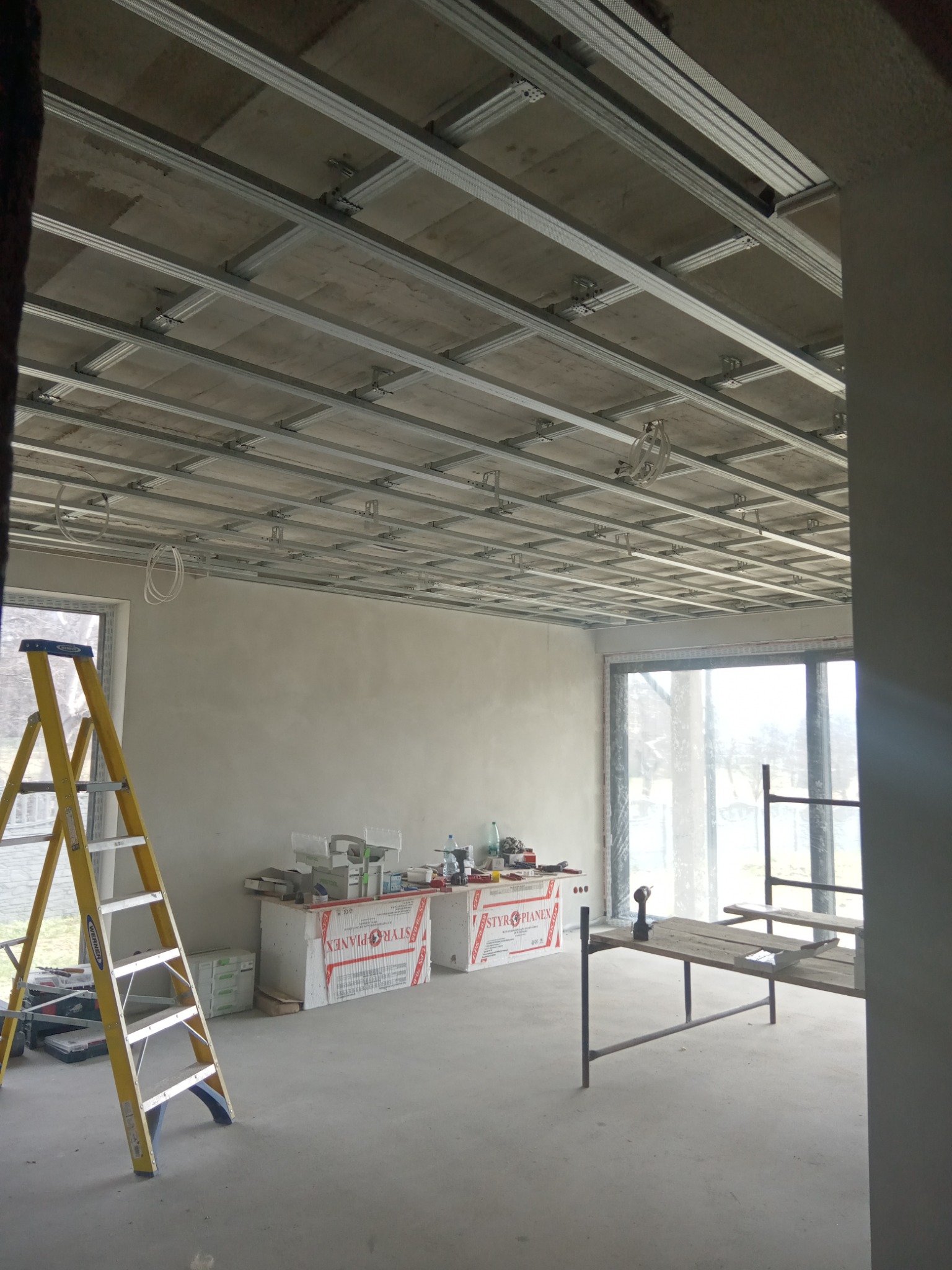 Stelaż sufitowy z metalowych profili w trakcie montażu, widoczne przewody elektryczne, żółta drabina, płyty styropianowe w tle, surowy stan wykończenia wnętrza.