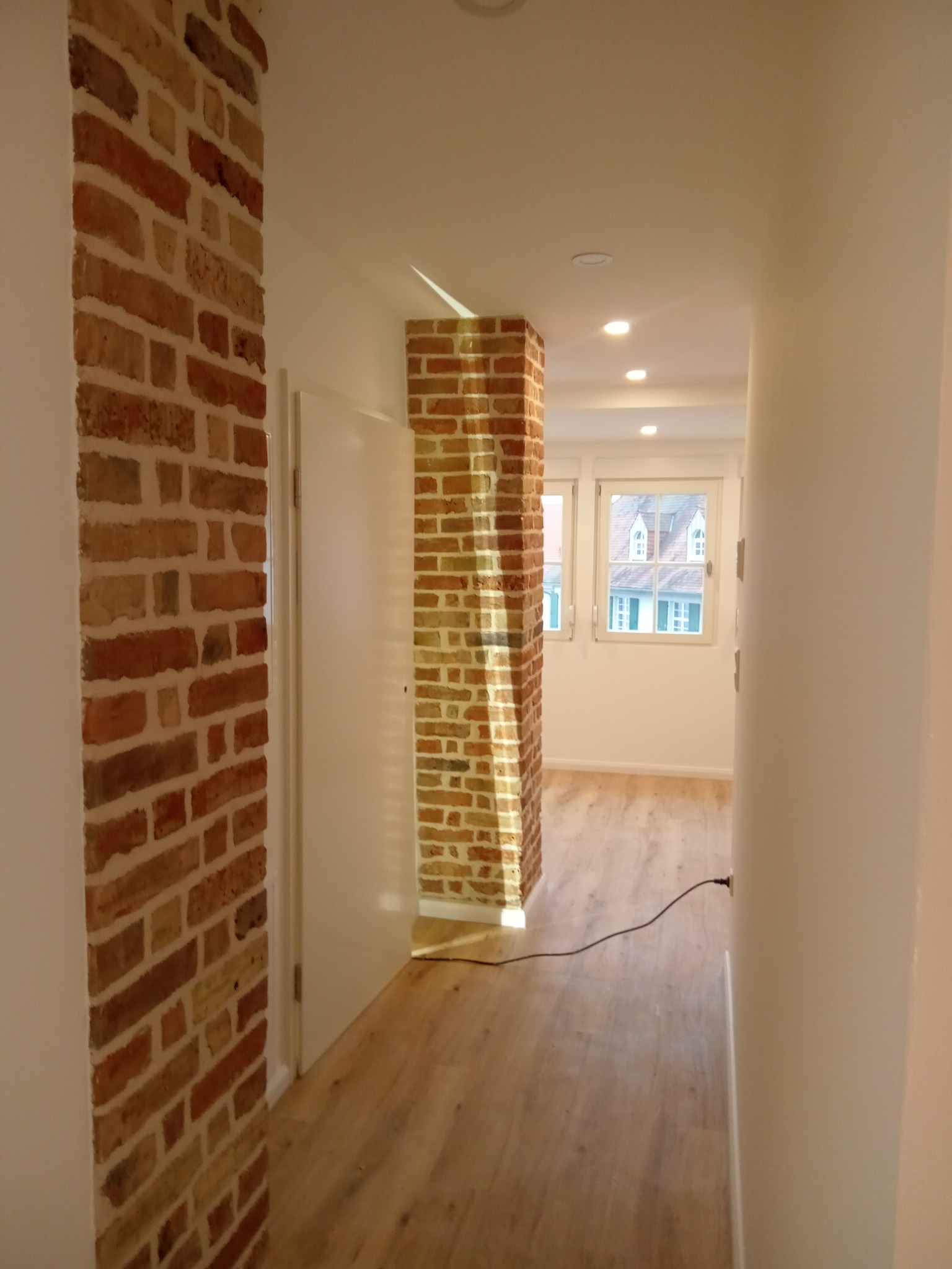 Wnętrze korytarza z wyeksponowanymi fragmentami muru z cegły, drewniana podłoga, białe ściany i drzwi, widok na jasne pomieszczenie z oknami.