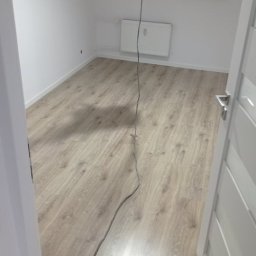 Łukasz Przespolewski - Pokój po remoncie: świeżo ułożone panele podłogowe w odcieniach szarości, białe ściany, grzejnik pod oknem z żaluzjami, widoczny kabel od urządzenia malarskiego.