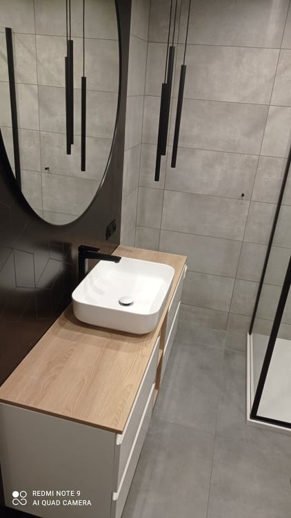 Nowoczesna łazienka z owalnym lustrem, wiszącymi lampami w kształcie czarnych prętów, białą umywalką nablatową na szafce z drewnianym blatem i szarą płytką podłogową.