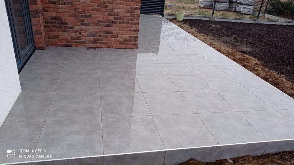 Szare płytki tarasowe ułożone na zewnątrz budynku z ceglaną ścianą i rabatą ziemi w tle.