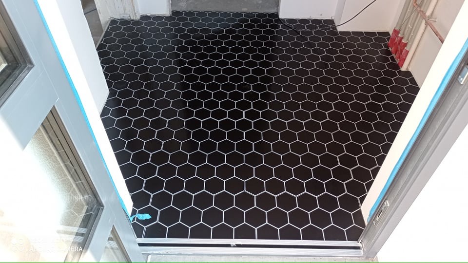 Ułożona czarna, heksagonalna podłoga z białymi fugami, widok z progu drzwi, z widocznymi rurami przy ścianie.