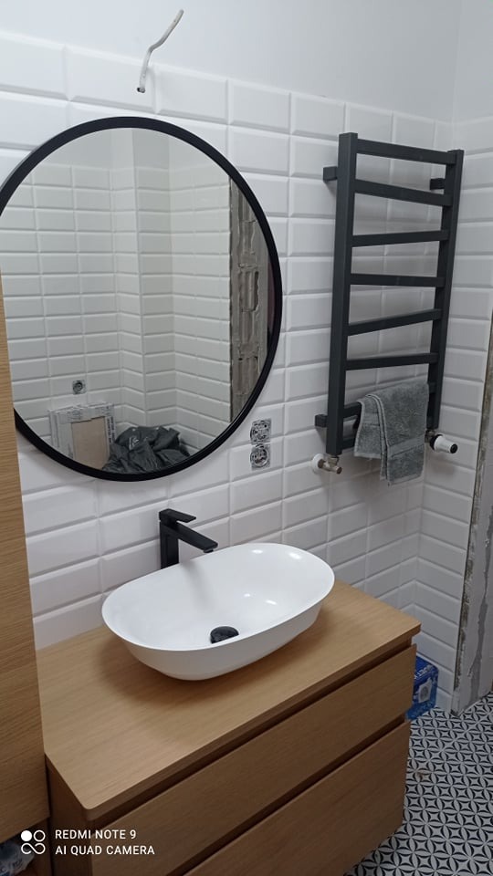 Nowoczesna łazienka z owalną umywalką nablatową, czarną armaturą, okrągłym lustrem w czarnej ramie i grzejnikiem drabinkowym w kolorze grafitowym na ścianie z białych płytek typu metro.