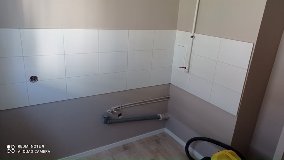 Świeżo wykafelkowana, jasna ściana w łazience z widocznymi rurami odpływowymi i instalacją elektryczną, przygotowana do dalszych prac wykończeniowych.