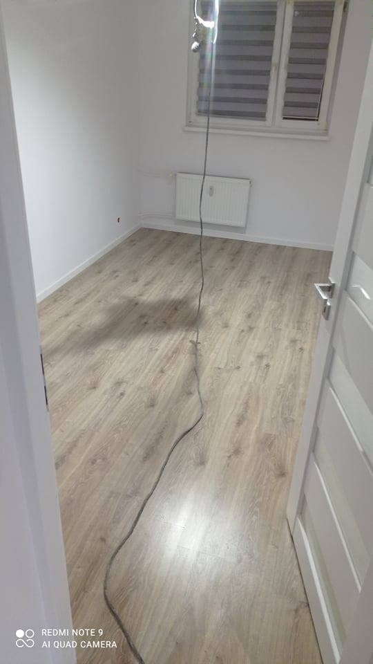 Pokój po remoncie: świeżo ułożone panele podłogowe w odcieniach szarości, białe ściany, grzejnik pod oknem z żaluzjami, widoczny kabel od urządzenia malarskiego.