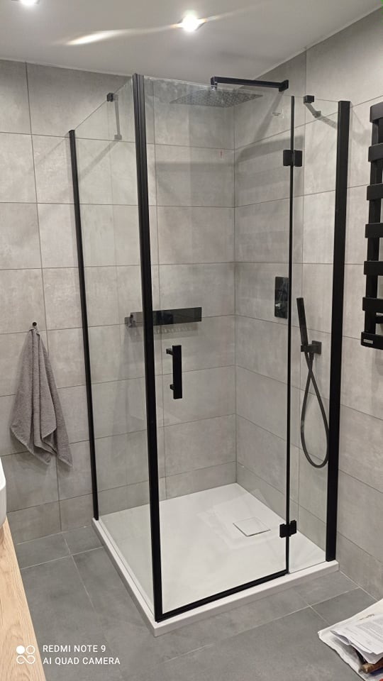 Nowoczesna kabina prysznicowa z czarnymi profilami i deszczownicą, zamontowana na białym brodziku w łazience wykończonej szarą płytką.