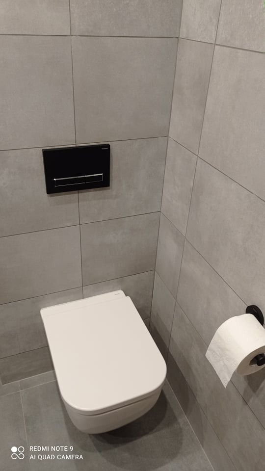 Nowoczesna toaleta podwieszana z czarnym panelem sterującym spłukiwaniem, obok uchwyt na papier toaletowy, szare płytki ścienne i podłogowe.