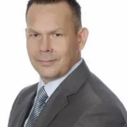 Portret mężczyzny w szarym garniturze i krawacie w paski na białym tle, uśmiechający się delikatnie.