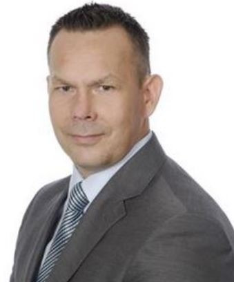 Portret mężczyzny w szarym garniturze i krawacie w paski na białym tle, uśmiechający się delikatnie.