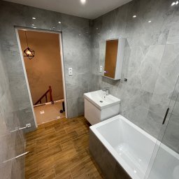 Maciej Wiśniewski Łazienki - Wykończona łazienka z szarymi płytkami imitującymi marmur, drewnopodobną podłogą i nowoczesną armaturą. Widok na schody przez otwarte drzwi.