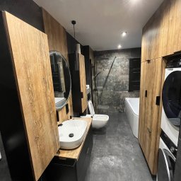 Maciej Wiśniewski Łazienki - Nowoczesna łazienka z drewnianymi akcentami: szafki, umywalka z okrągłym lustrem, toaleta, wanna i pralka. Szare płytki podłogowe i ściany w strefie prysznica.