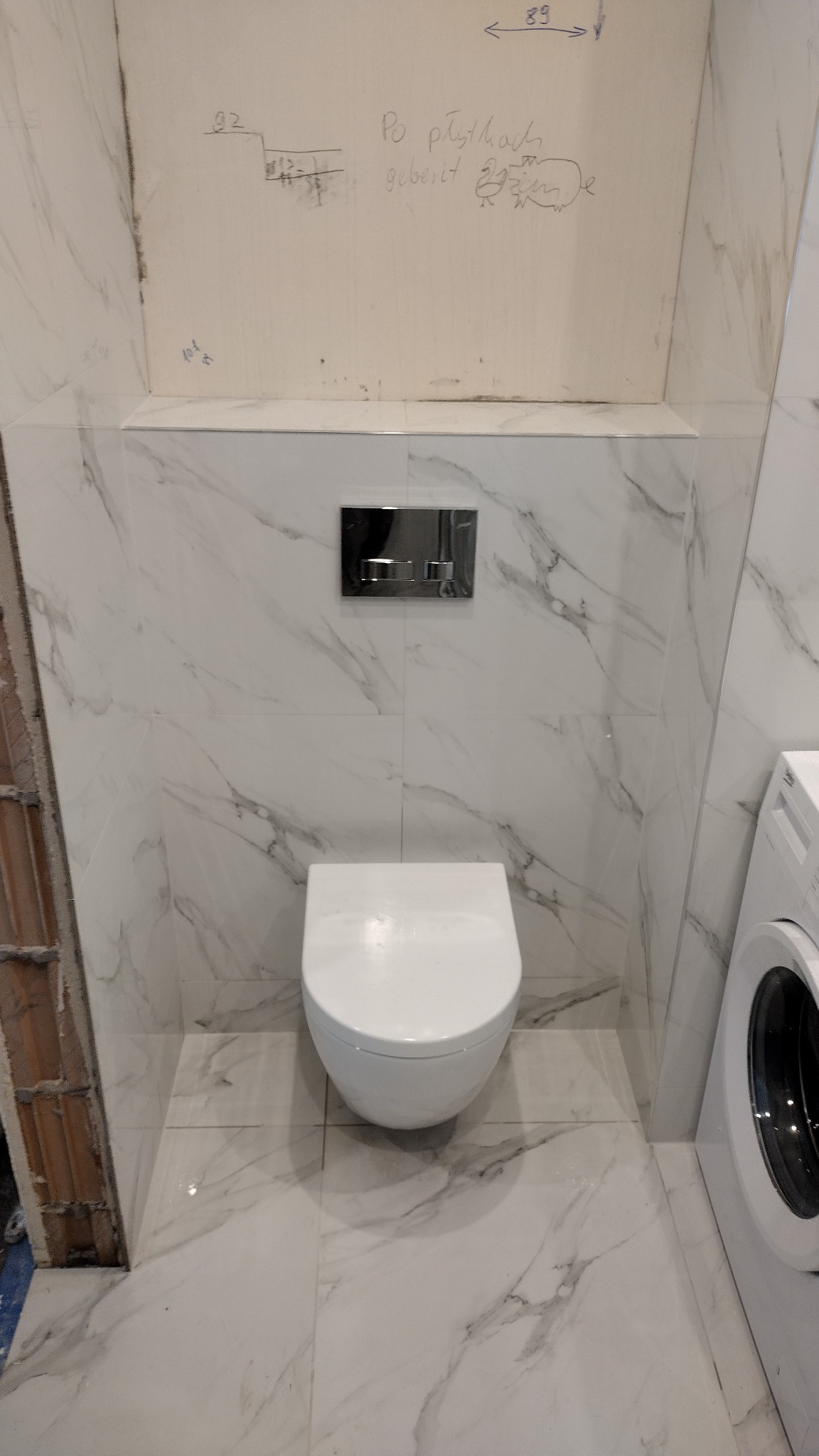 Podwieszana toaleta w trakcie montażu, widoczne białe płytki imitujące marmur, ściana z niedokończonymi pracami i odręcznymi notatkami, obok pralka.