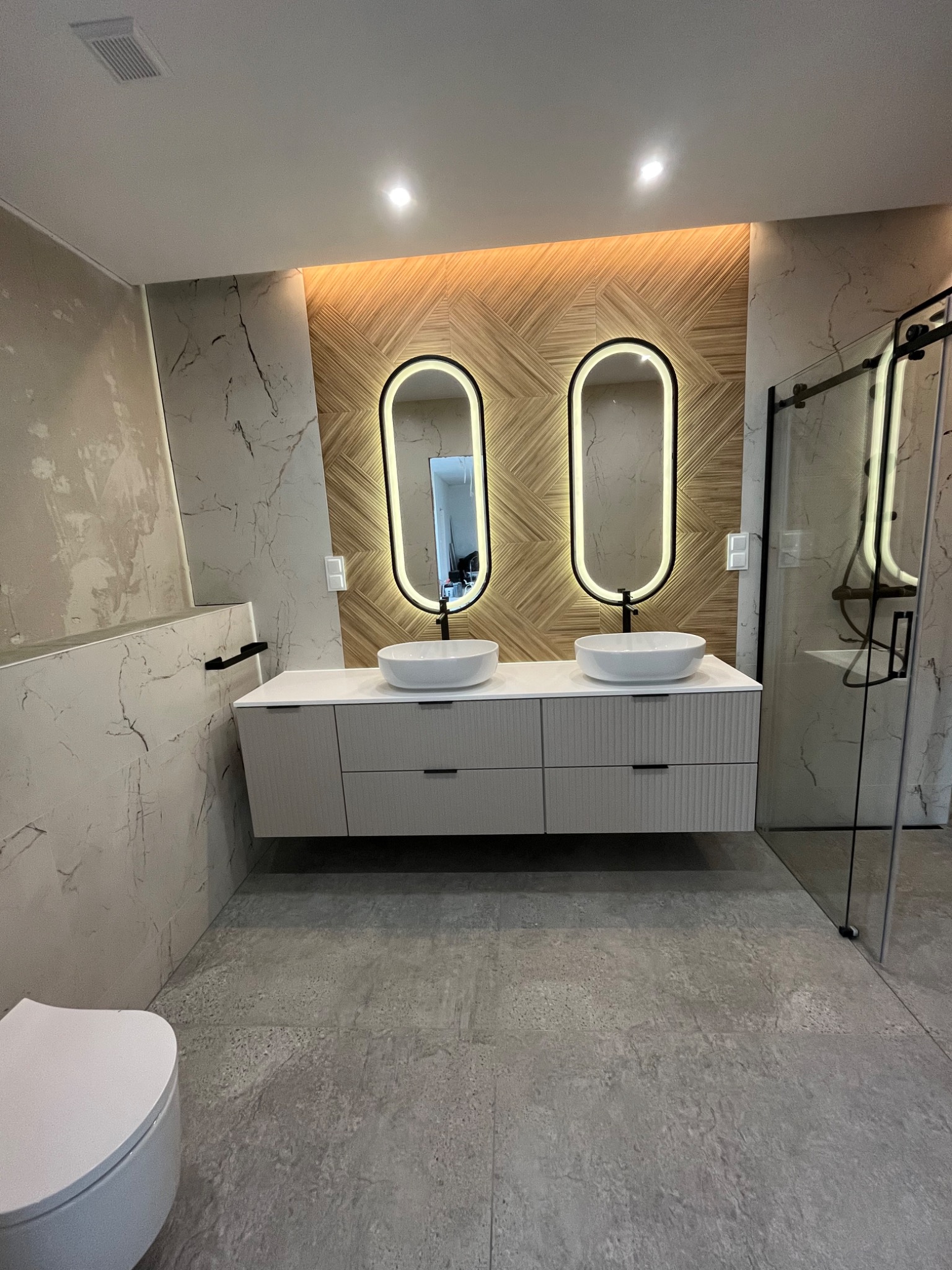 Nowoczesna łazienka z dwoma umywalkami nablatowymi, podświetlanymi lustrami w owalnych ramach i kabiną prysznicową ze szklanymi drzwiami. Ściany wykończone płytkami imitującymi marmur.