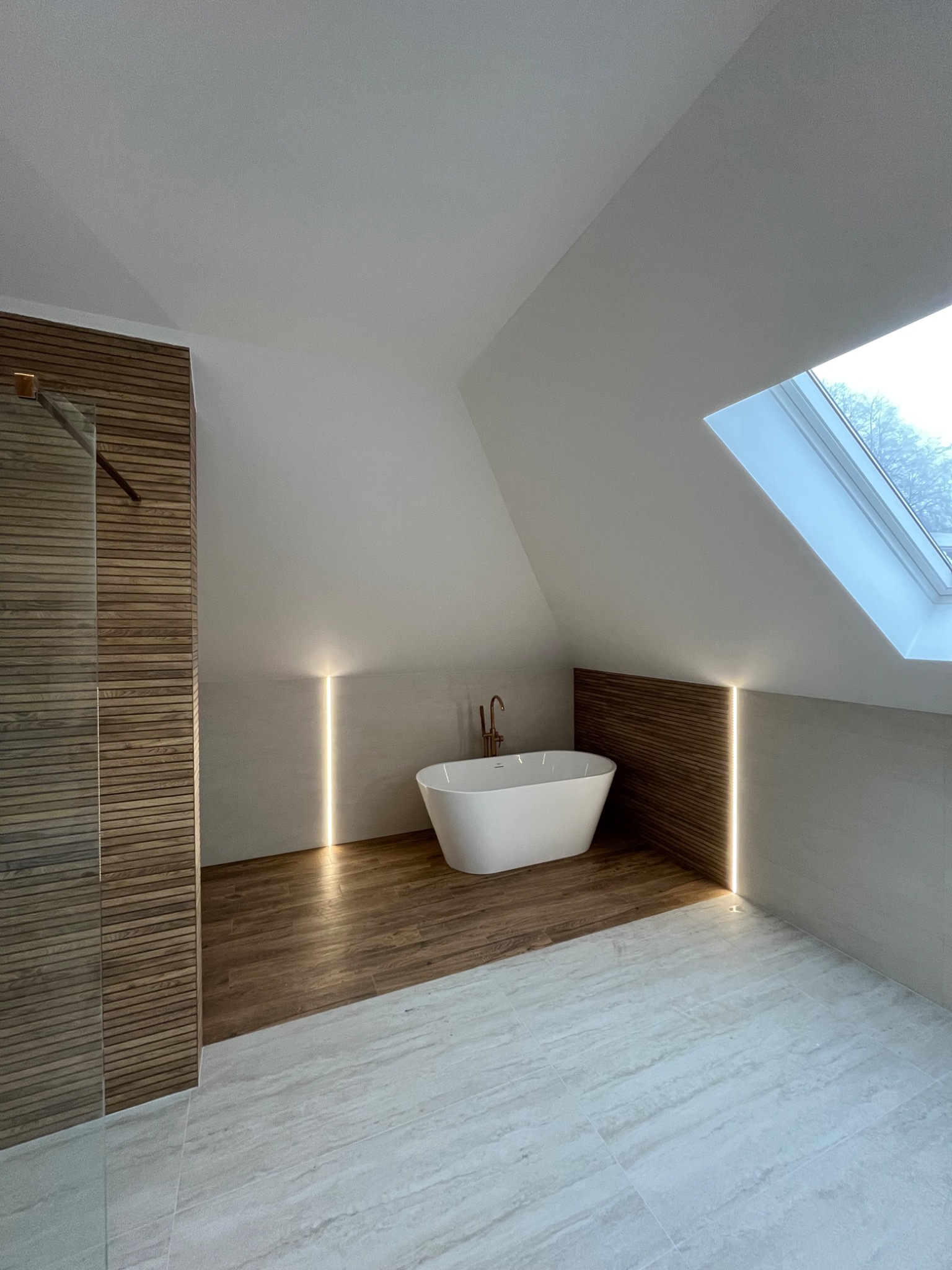 Nowoczesna łazienka na poddaszu z wolnostojącą wanną, drewnianą okładziną i subtelnym oświetleniem LED. Uwagę przykuwa okno dachowe i marmurowe płytki podłogowe.