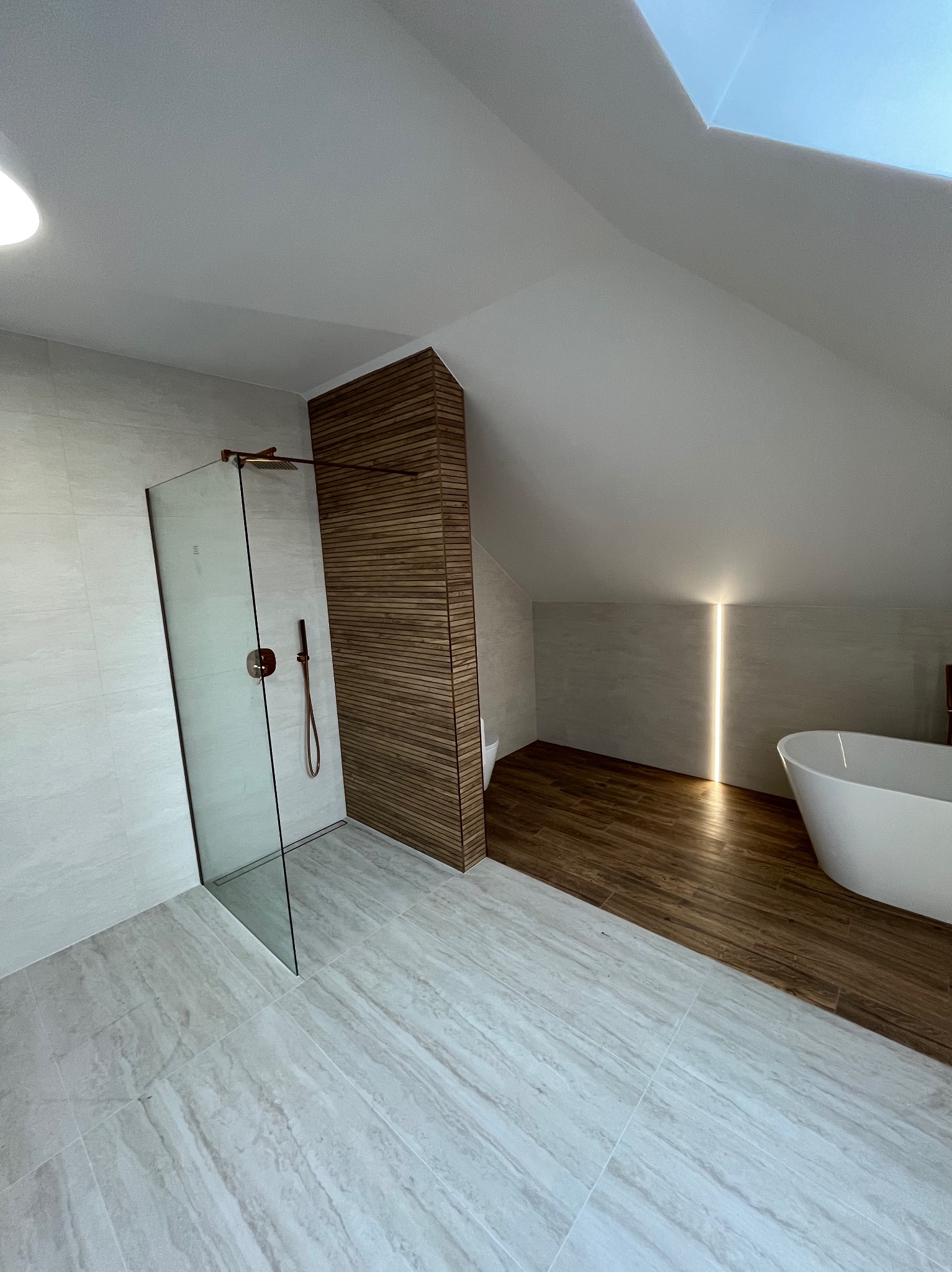 Nowoczesna łazienka na poddaszu z kabiną prysznicową ze szklaną ścianką, drewnianą ścianką działową i wolnostojącą wanną. Uwagę przykuwa oświetlenie LED w ścianie.