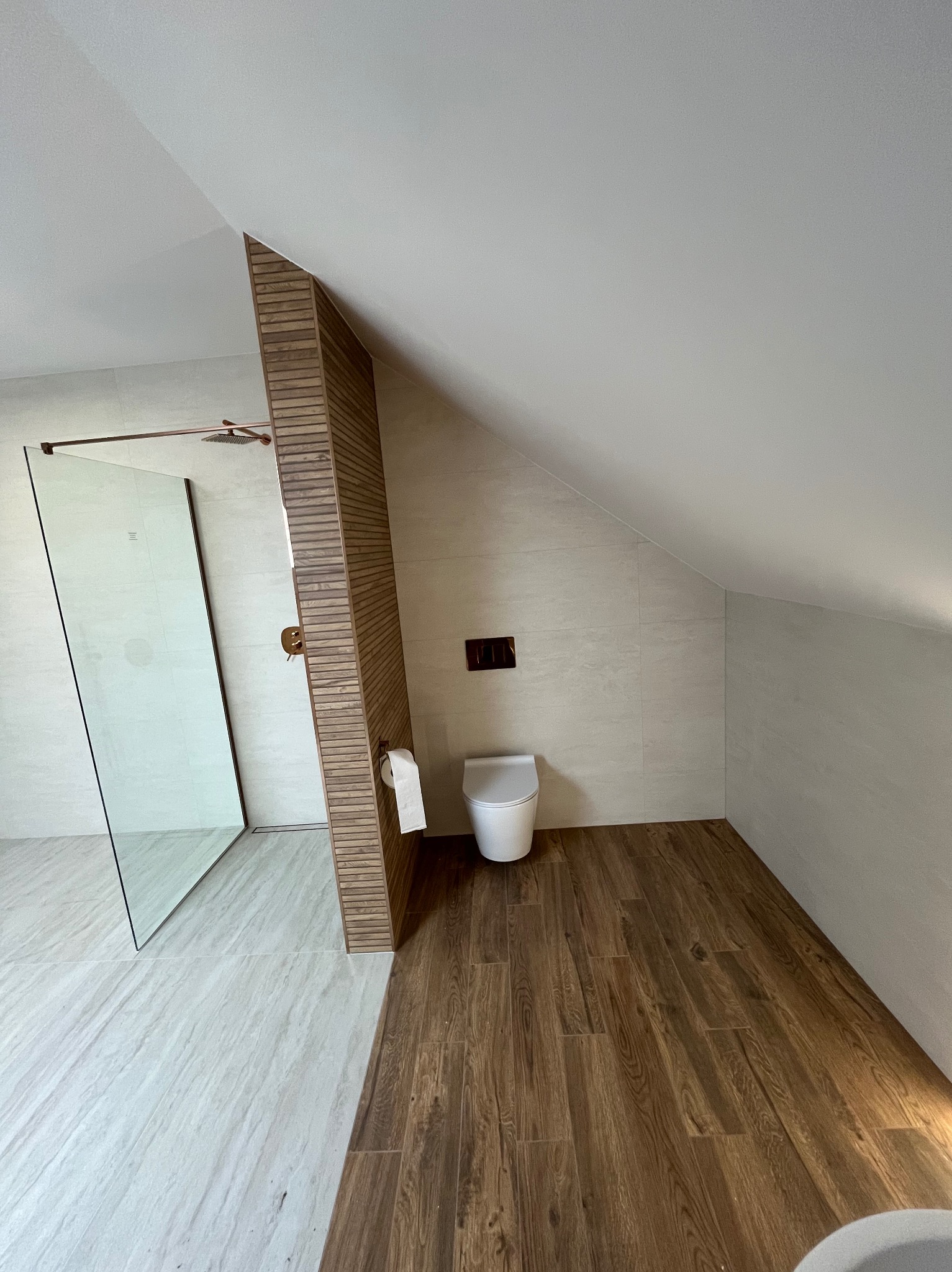 Nowoczesna łazienka na poddaszu z kabiną prysznicową typu walk-in, toaletą i drewnianą ścianką działową. Jasne płytki na ścianach i podłodze, drewniana podłoga.