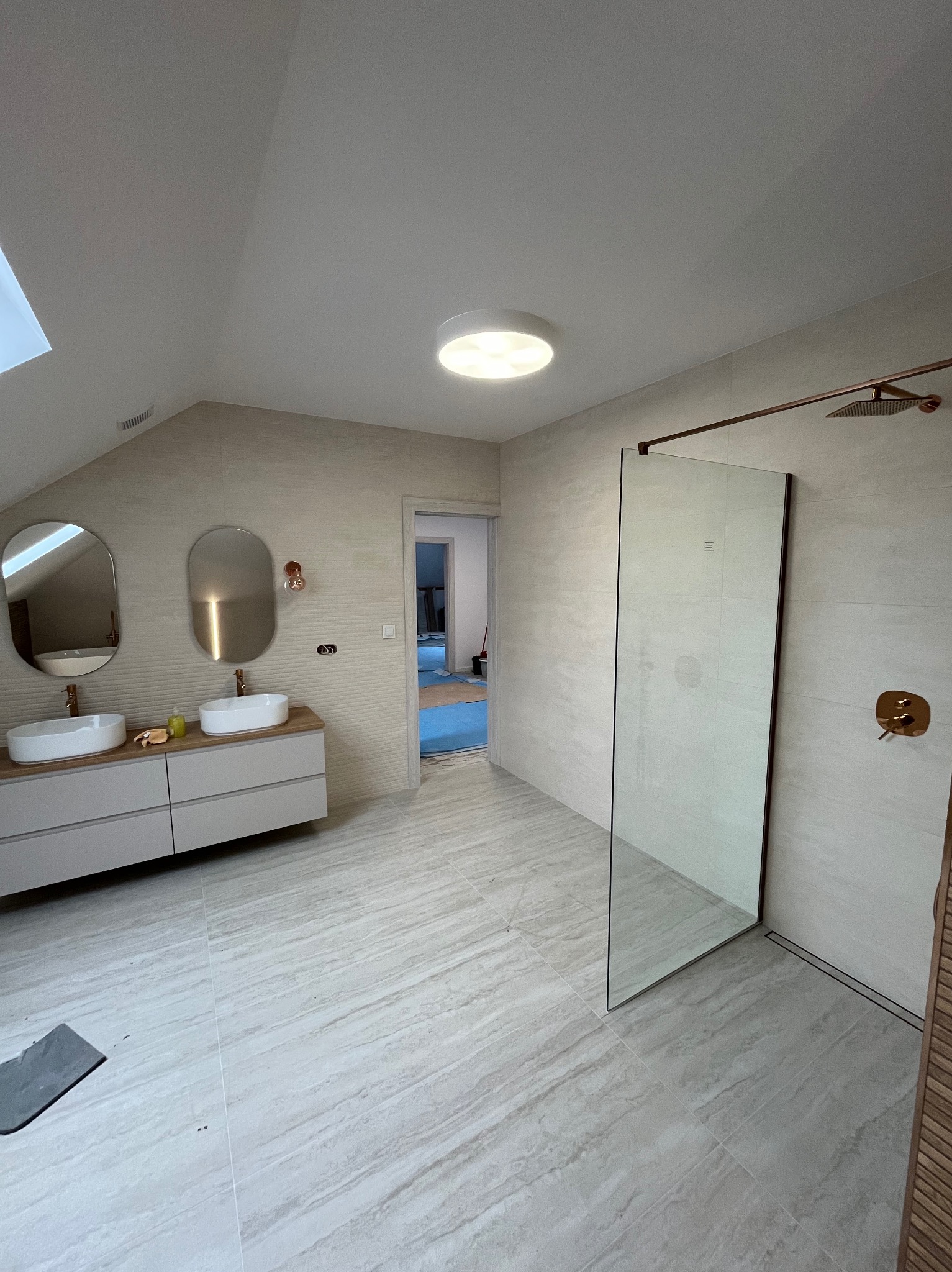 Jasna łazienka na poddaszu z dwoma umywalkami, kabiną prysznicową typu walk-in z miedzianymi elementami i oświetleniem sufitowym. Widoczne płytki imitujące kamień.