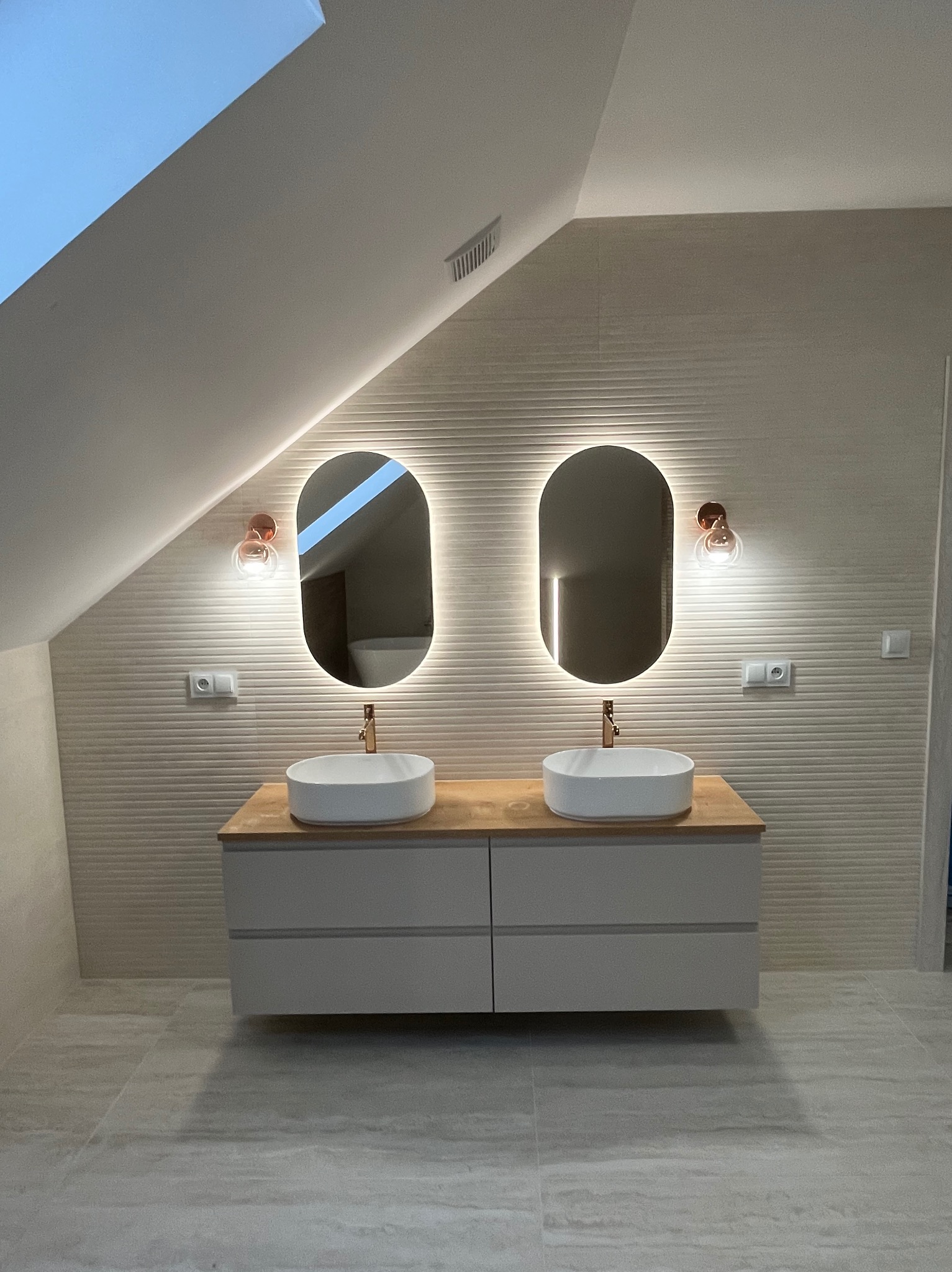 Nowoczesna łazienka na poddaszu z dwoma umywalkami, podświetlanymi lustrami i beżowymi płytkami z poziomym wzorem. Umywalki owalne, białe, z bateriami w kolorze miedzi.