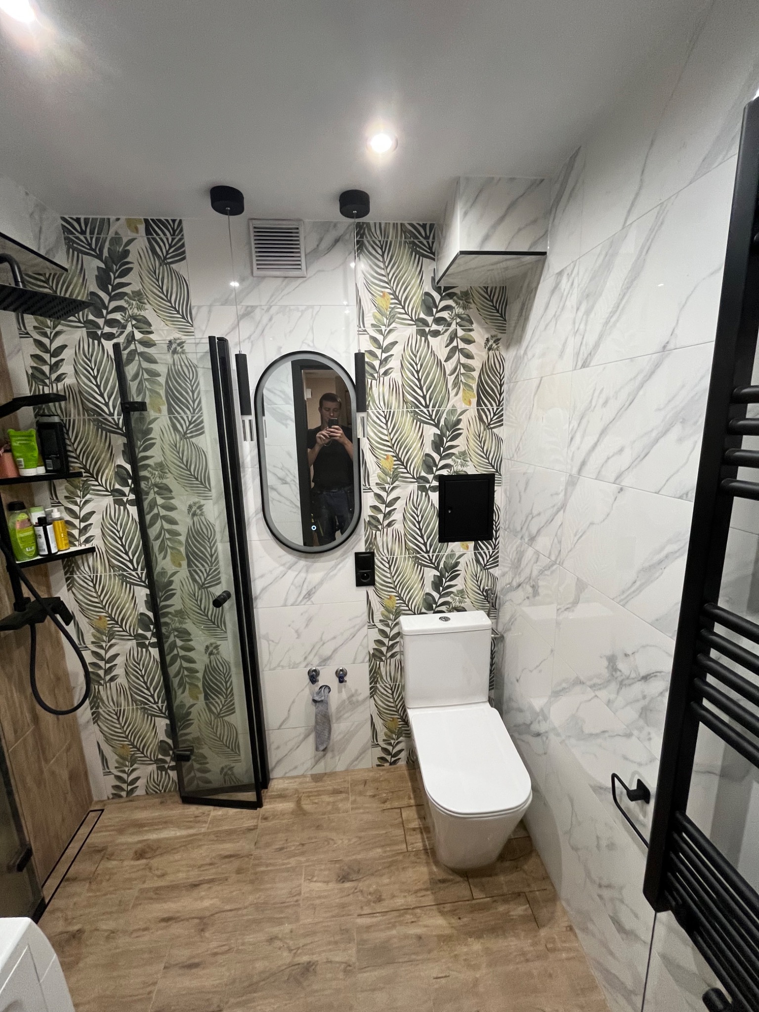 Nowoczesna łazienka z kabiną prysznicową typu walk-in, toaletą i owalnym lustrem. Ściany wyłożone płytkami imitującymi marmur i z motywem liści. Czarny grzejnik drabinkowy.