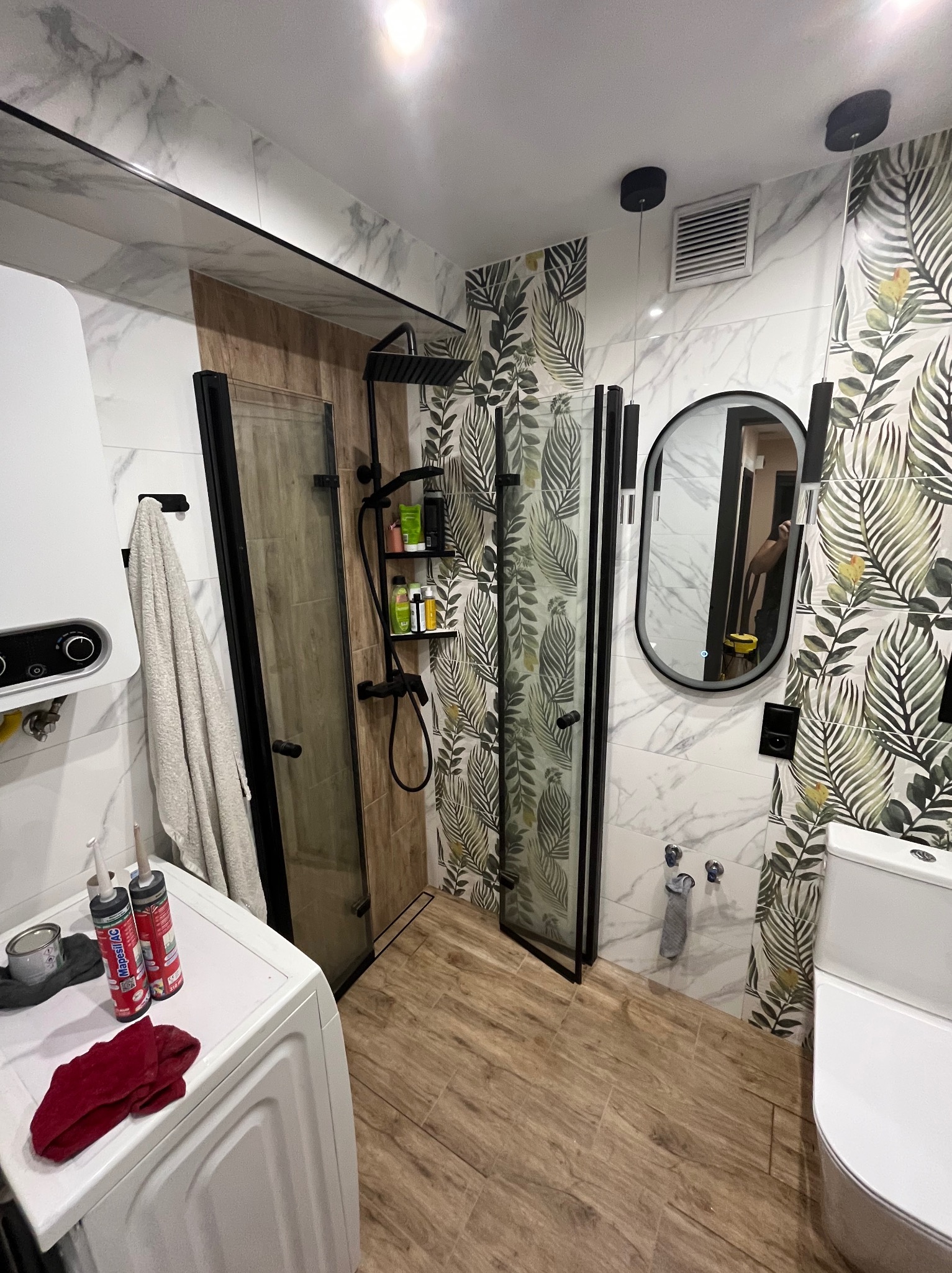 Nowoczesna łazienka z kabiną prysznicową typu walk-in, marmurowymi i roślinnymi płytkami, drewnopodobną podłogą oraz czarnymi akcentami.