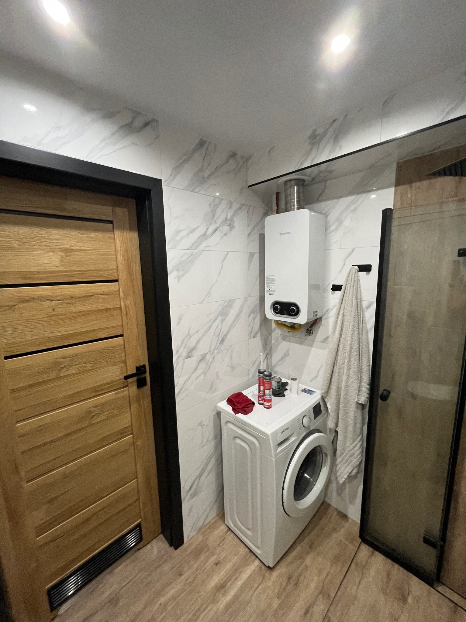 Wykończona łazienka z pralką, piecem gazowym i kabiną prysznicową. Ściany wyłożone płytkami imitującymi marmur, drewniane drzwi z czarną ościeżnicą.