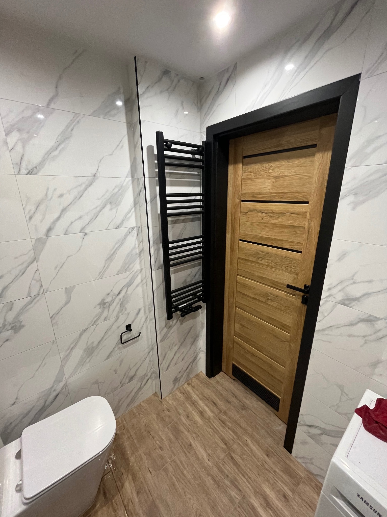Nowoczesna łazienka z marmurowymi płytkami, drewnianą podłogą i drzwiami z czarną ościeżnicą. Grzejnik drabinkowy w kolorze czarnym. Minimalistyczny design.