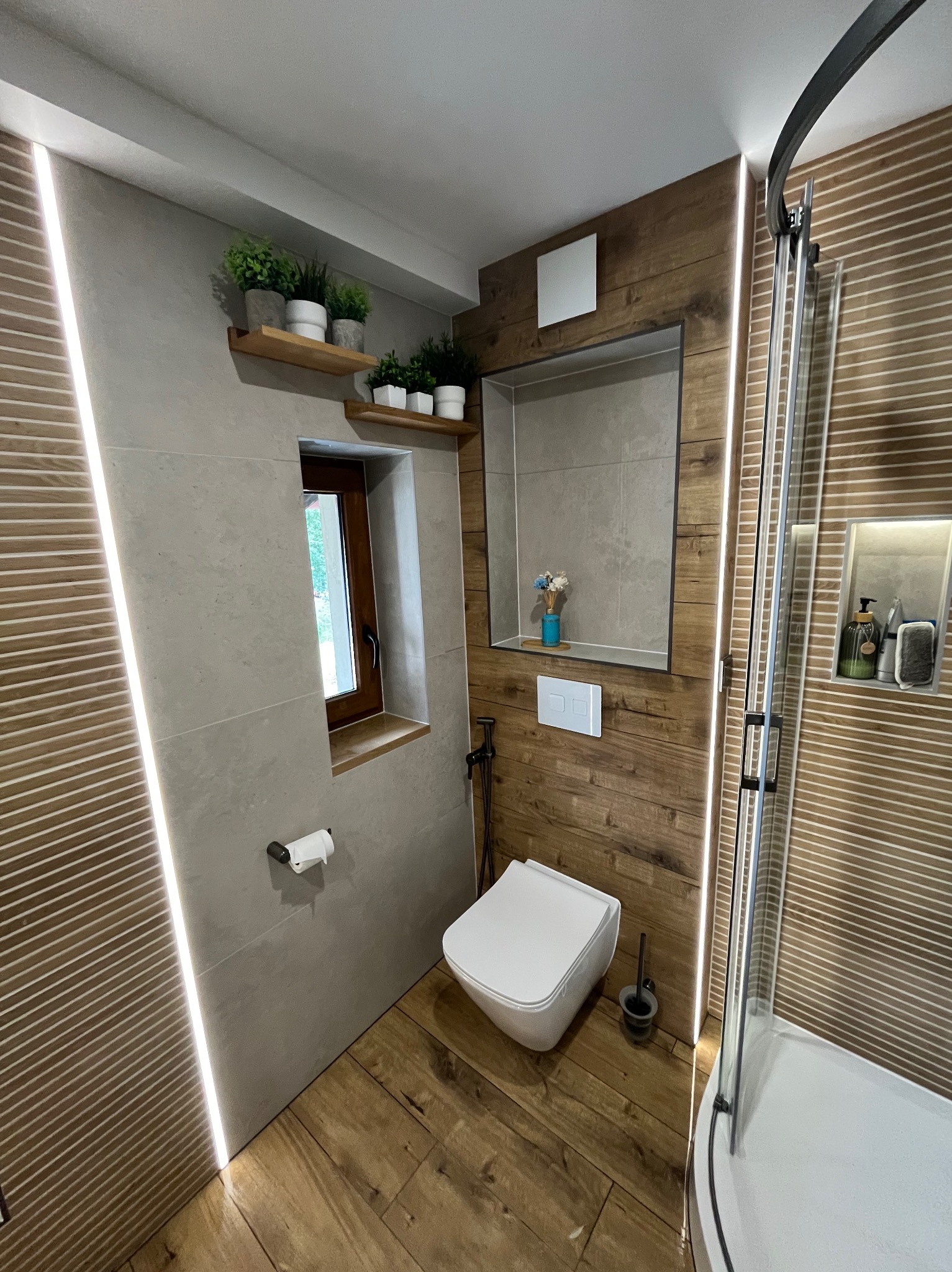 Nowoczesna łazienka po remoncie: drewnopodobne płytki, szara ściana z oknem i półkami z roślinami, kabina prysznicowa z czarnymi detalami, oświetlenie LED.