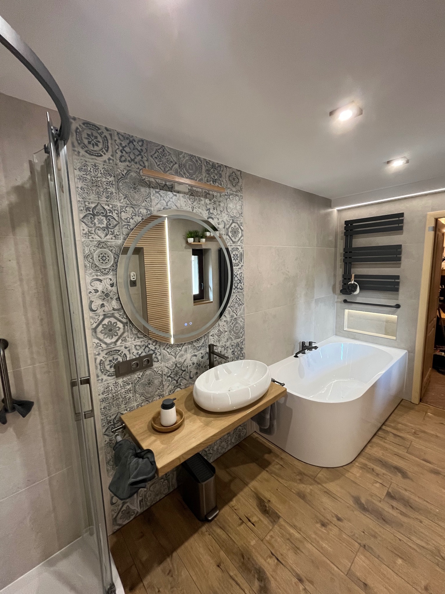 Nowoczesna łazienka z okrągłym lustrem LED, drewnianym blatem pod umywalką i wanną. Dekoracyjne płytki na ścianie, drewnopodobna podłoga. Minimalistyczny grzejnik.