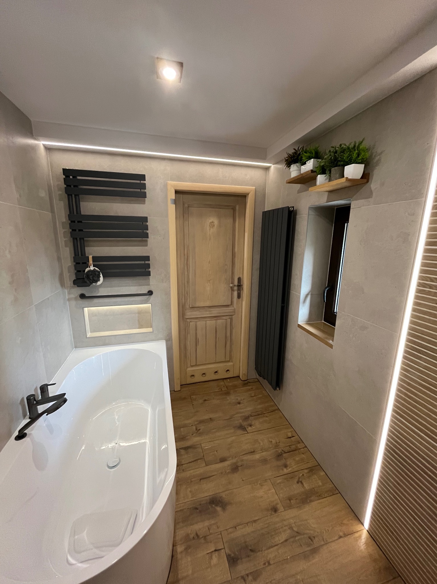 Nowoczesna łazienka z drewnianą podłogą i akcentami, białą wanną, grzejnikiem drabinkowym i roślinami na półce przy oknie. Oświetlenie LED podkreśla detale.