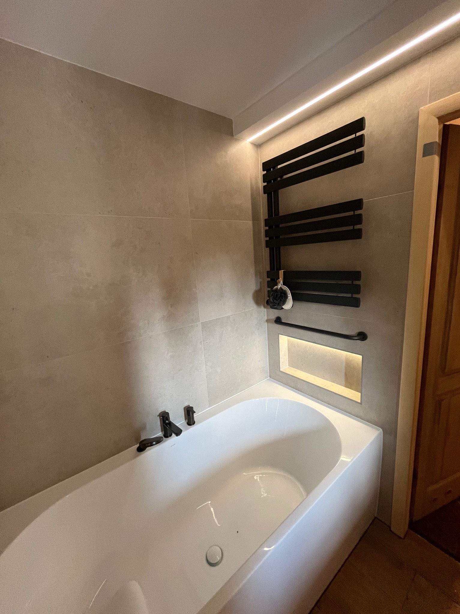 Nowoczesna łazienka z beżowymi płytkami, białą wanną i czarnym grzejnikiem drabinkowym. Podświetlana wnęka dodaje ciepła. Minimalistyczny design.