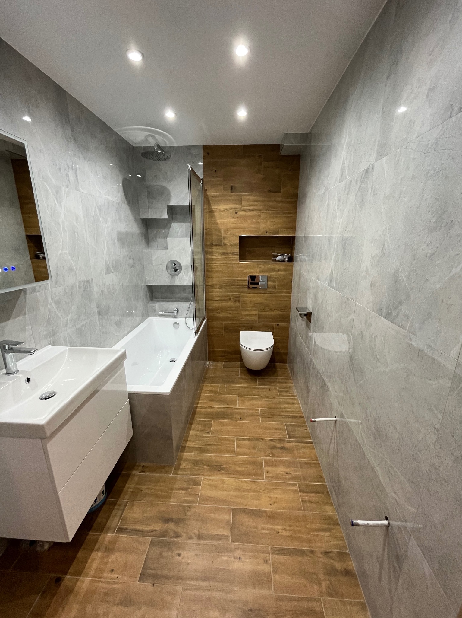 Nowoczesna łazienka z szarymi płytkami imitującymi marmur i drewnopodobną podłogą. Umywalka z szafką, wanna z parawanem, toaleta podtynkowa i wnęki w ścianie.