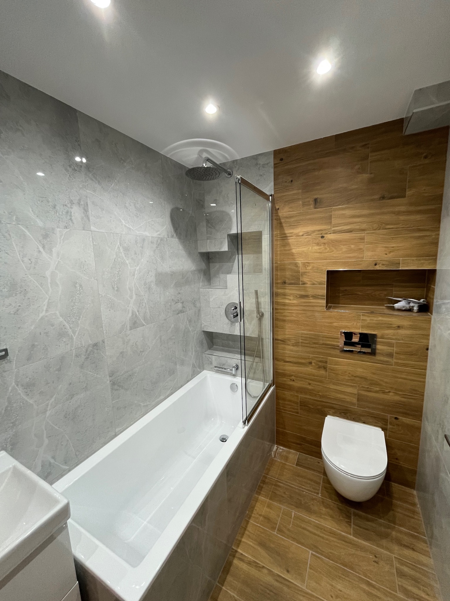 Nowoczesna łazienka z wanną za szklanym parawanem, szare płytki imitujące marmur i drewnopodobne płytki na podłodze i ścianie. W tle toaleta i wnęka w ścianie.