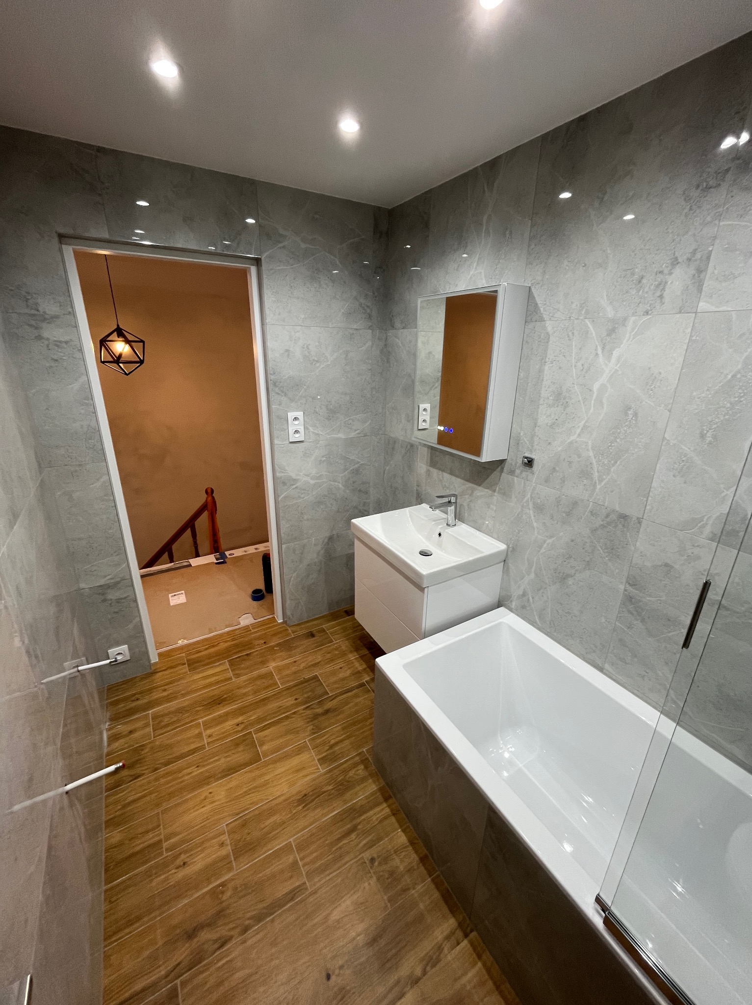 Wykończona łazienka z szarymi płytkami imitującymi marmur, drewnopodobną podłogą i nowoczesną armaturą. Widok na schody przez otwarte drzwi.