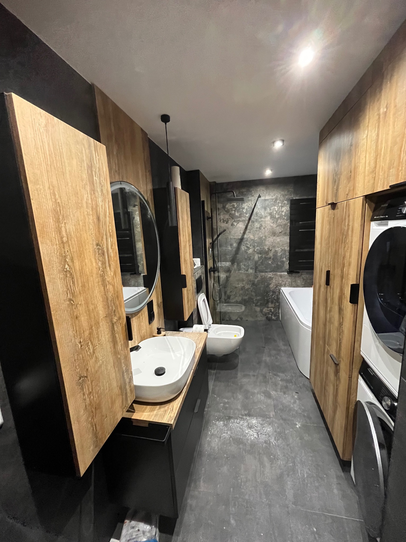 Nowoczesna łazienka z drewnianymi akcentami: szafki, umywalka z okrągłym lustrem, toaleta, wanna i pralka. Szare płytki podłogowe i ściany w strefie prysznica.