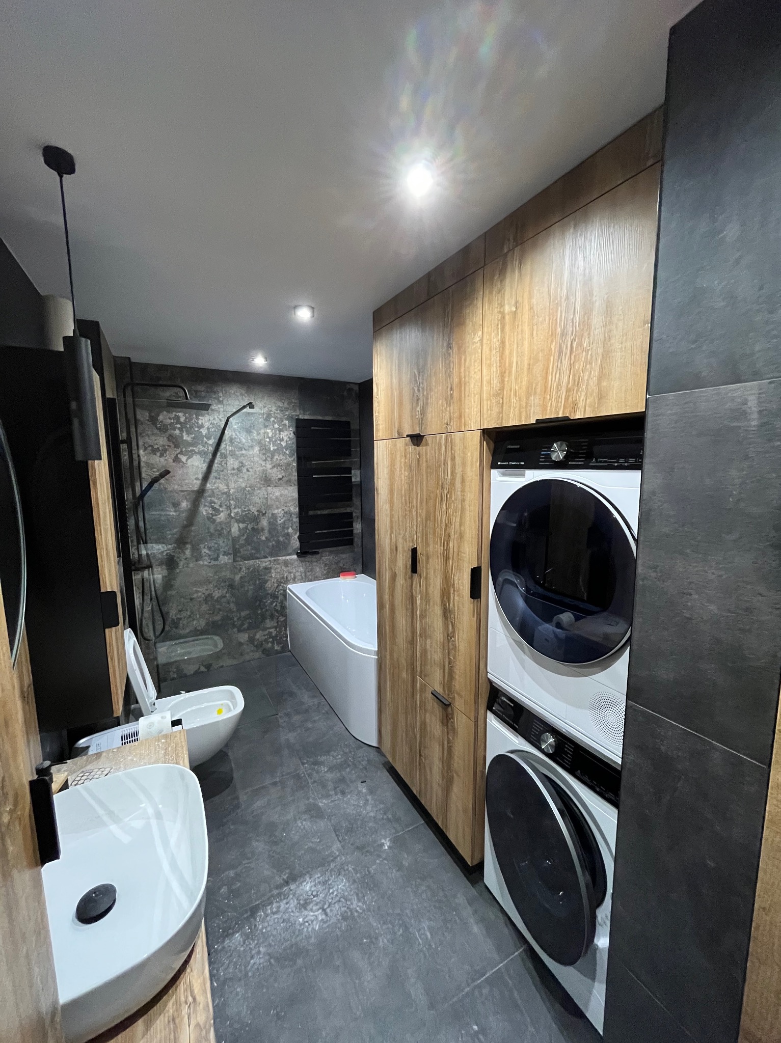 Nowoczesna łazienka z drewnianymi szafkami, pralko-suszarką w zabudowie, umywalką nablatową i kabiną prysznicową z deszczownicą. Szare płytki na ścianach i podłodze.