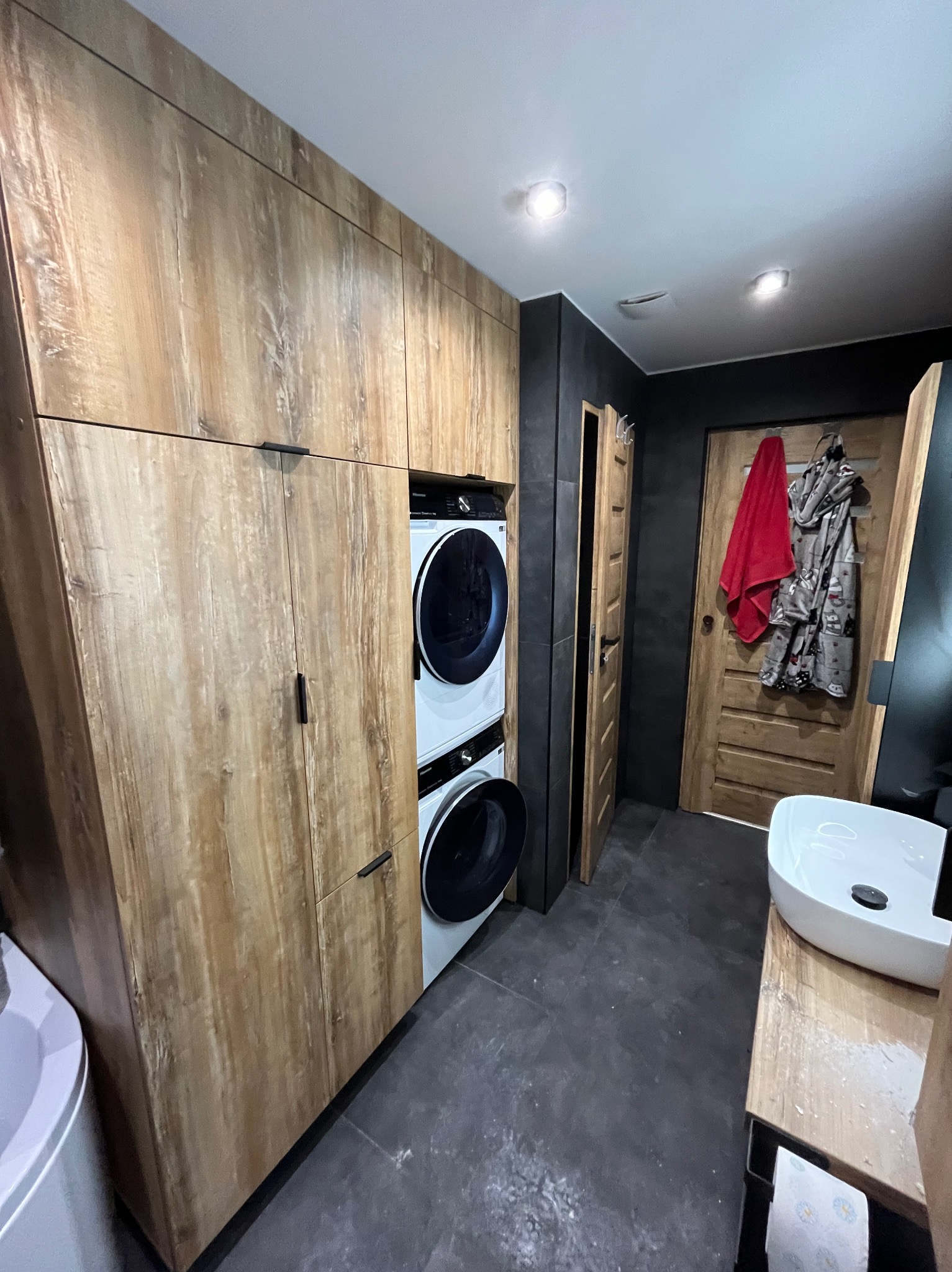 Nowoczesna łazienka z drewnianą zabudową, pralką i suszarką w kolumnie, umywalką nablatową i ciemnymi płytkami podłogowymi. Funkcjonalne i stylowe rozwiązanie.