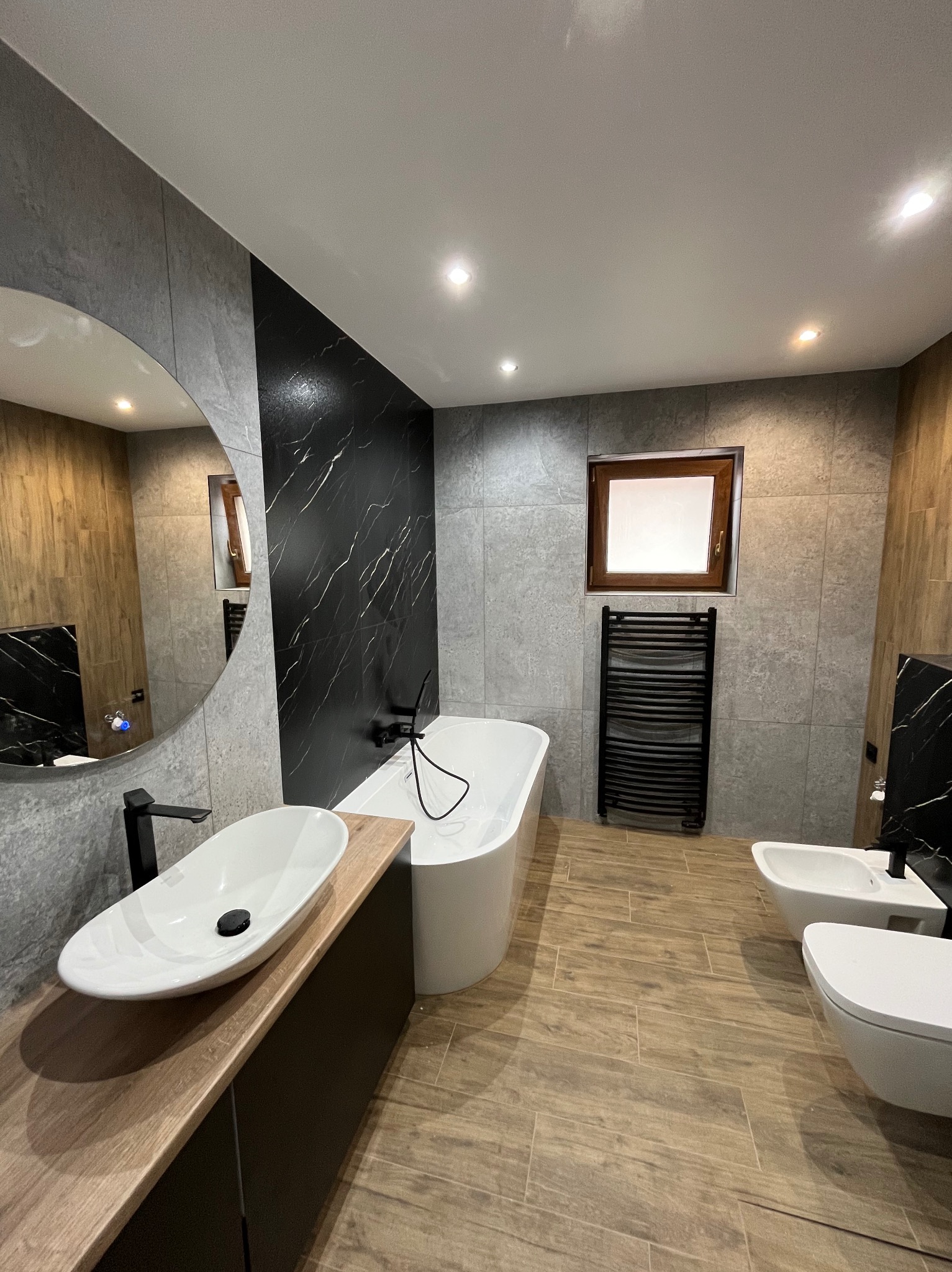 Nowoczesna łazienka z owalną umywalką na drewnianym blacie, białą wanną i czarnymi akcentami: bateria, grzejnik, ściana z marmuru. Jasne płytki i drewnopodobna podłoga.