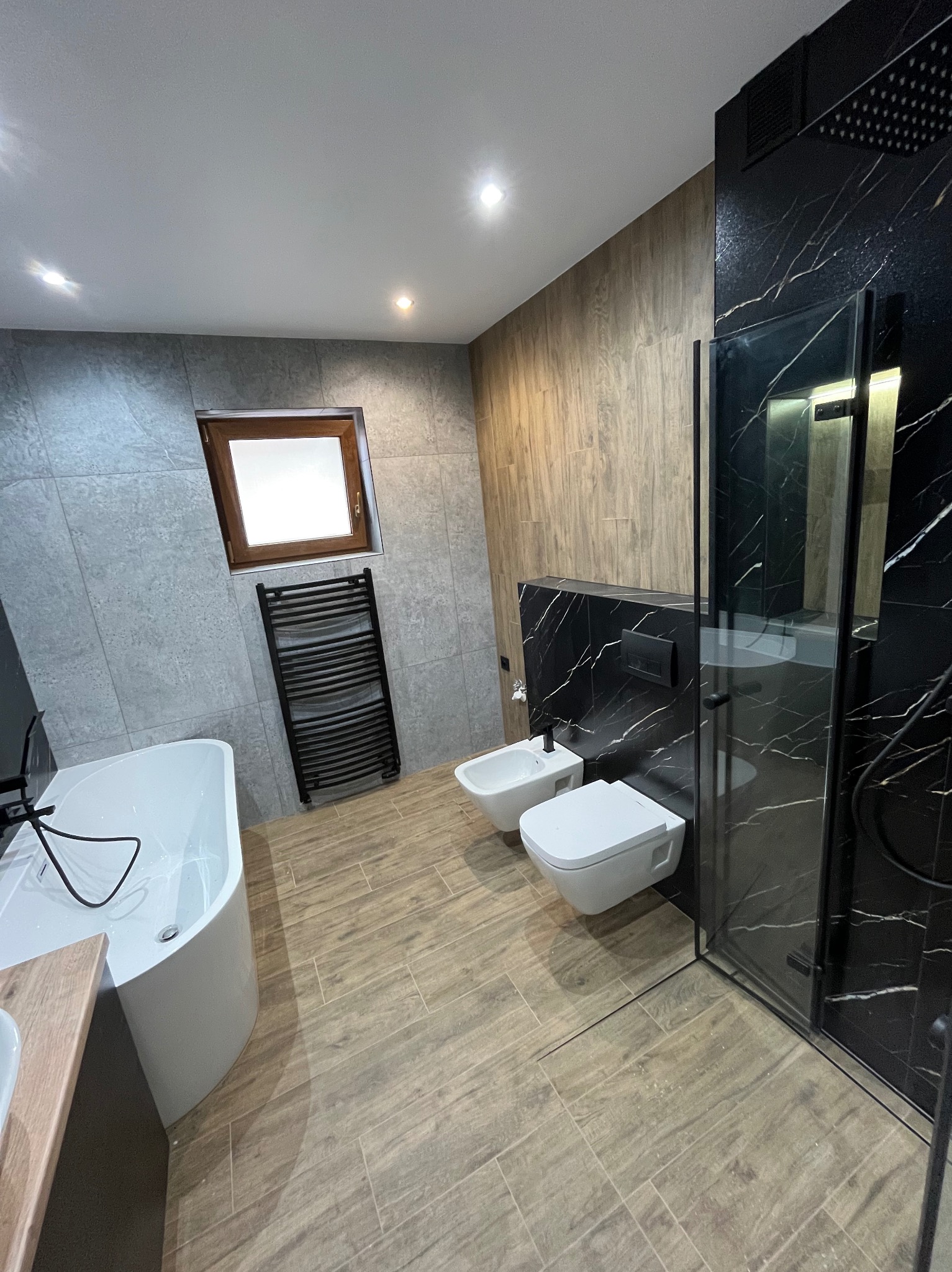 Nowoczesna łazienka z kabiną prysznicową typu walk-in, białą armaturą i grzejnikiem drabinkowym. Płytki imitujące drewno na podłodze i szare na ścianach.