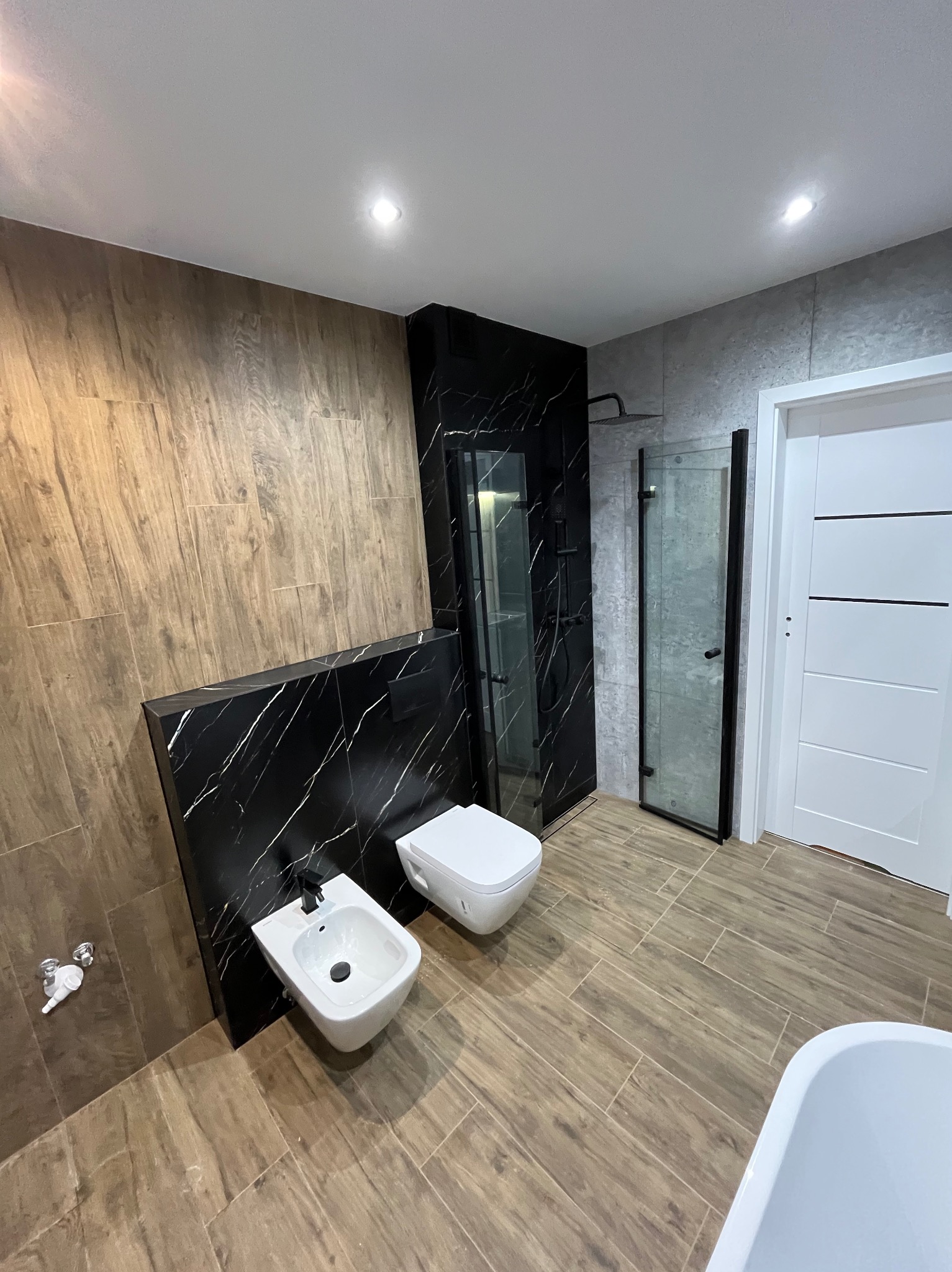 Nowoczesna łazienka z drewnopodobnymi płytkami, czarnym marmurem i kabiną prysznicową. Bidet i toaleta podwieszana. Oświetlenie punktowe.