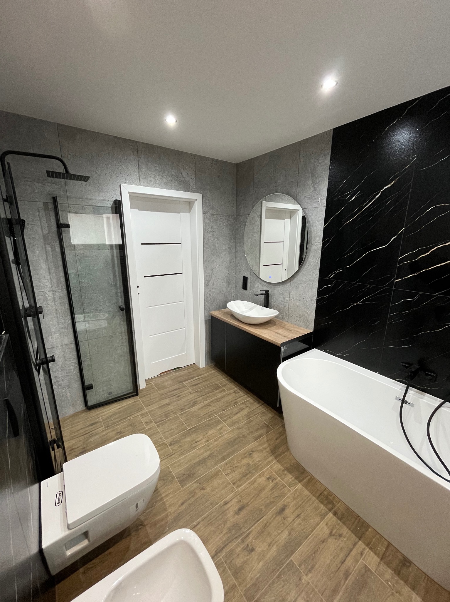 Nowoczesna łazienka z czarno-białymi akcentami: kabina prysznicowa typu walk-in, wolnostojąca wanna, umywalka nablatowa i płytki imitujące marmur.