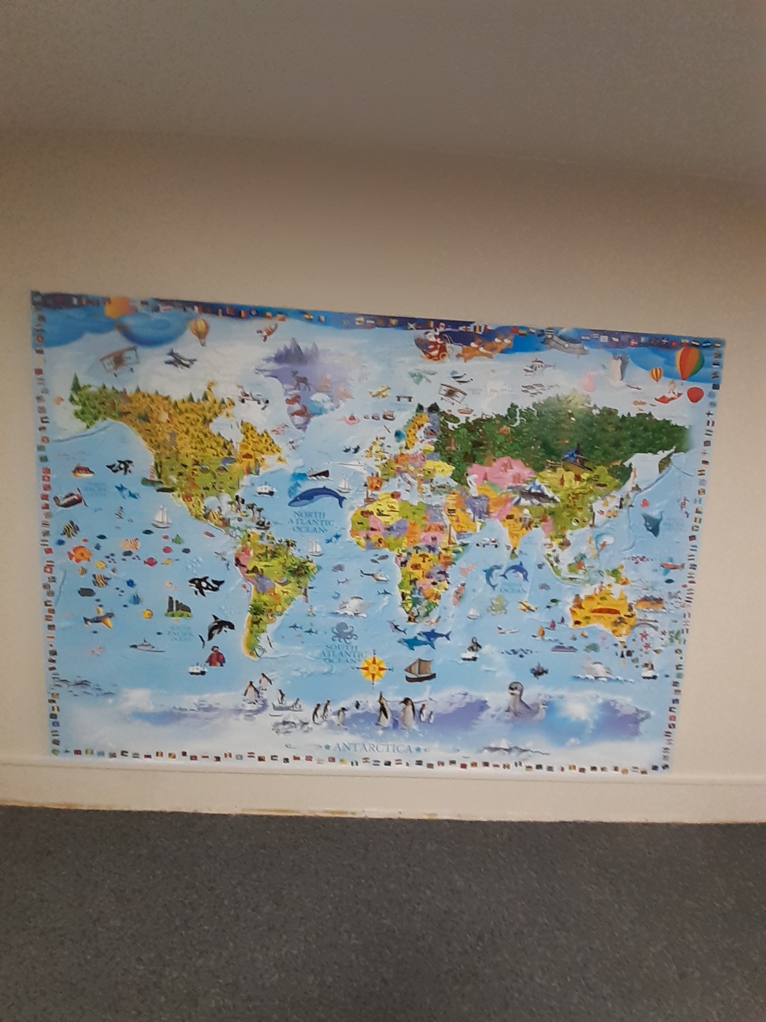Ilustrowana mapa świata z rysunkami zwierząt, statków i samolotów, wisząca na białej ścianie nad szarą wykładziną.