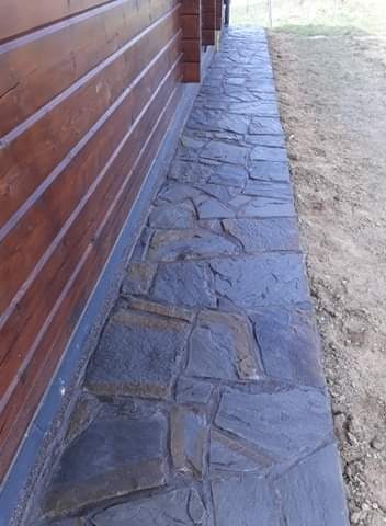 Ścieżka z nieregularnych, grafitowych płyt kamiennych przylegająca do drewnianej, poziomo ułożonej elewacji domu. Widoczny grunt obok ścieżki.