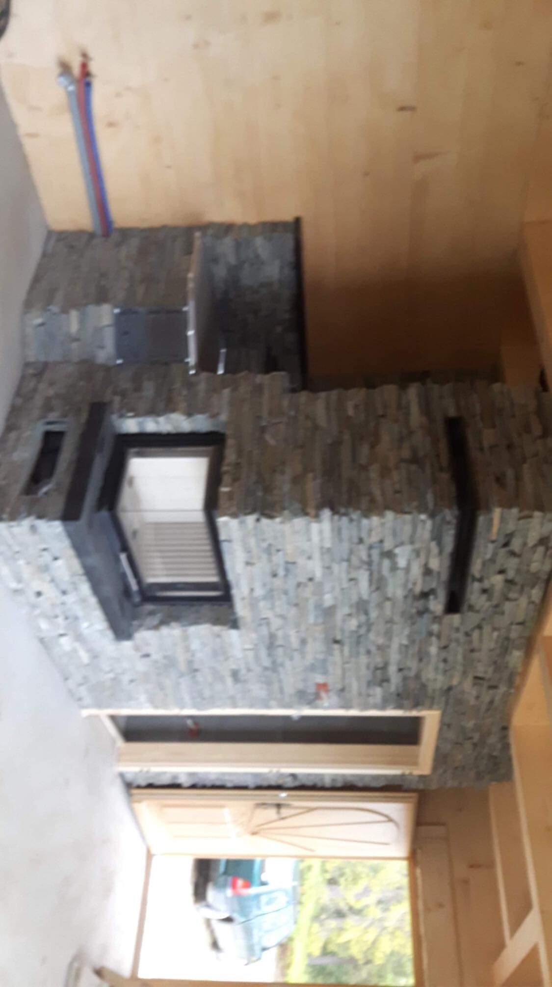 Wnętrze w Ostrowsku w trakcie wykańczania kominka obłożonego szarą, łupkową okładziną kamienną z czarnymi wstawkami, widoczne elementy konstrukcyjne i instalacja hydrauliczna.